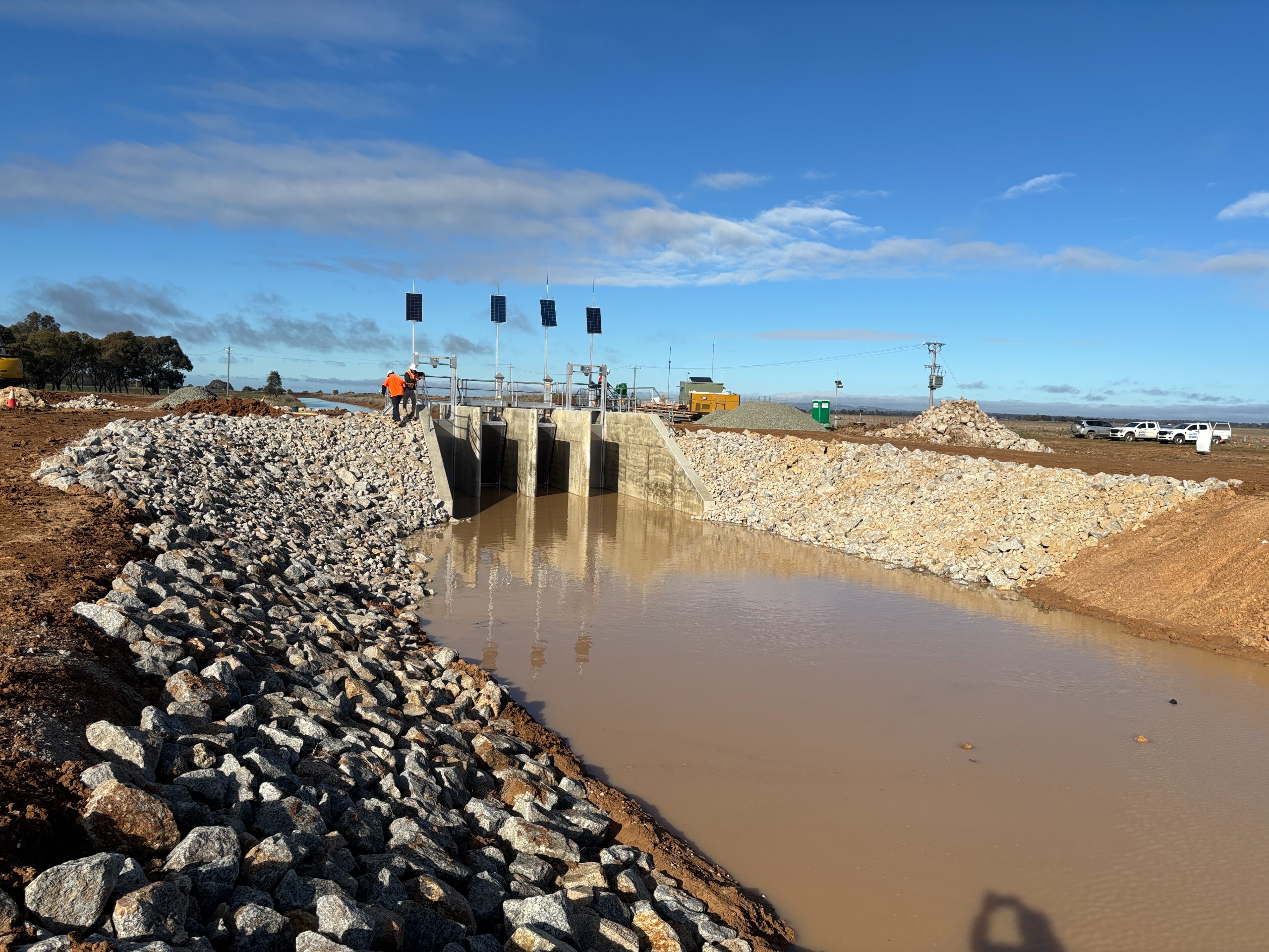 An image showing water flowing along a channel with blue sky, with a newly-constructed flow regulator in the background. 