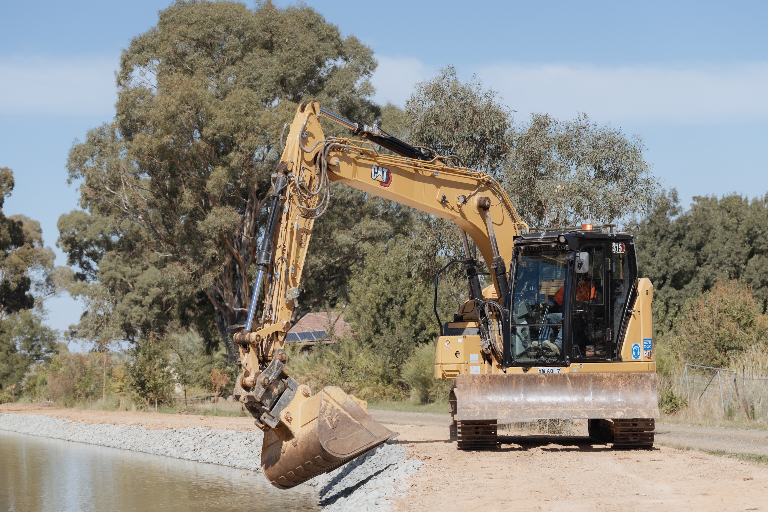 A yellow excavator working to remodel a water channel bank, with the bucket scooping alongside the channel wall. 
